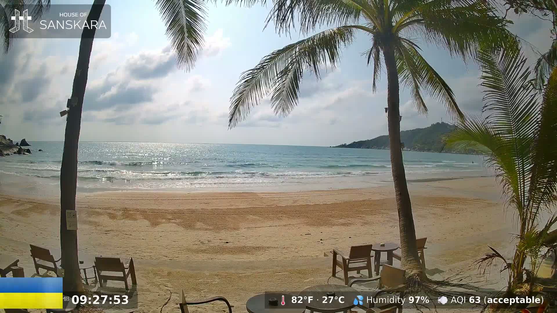 A mostly sunny beach scene shows calm ocean waves lapping a sandy shore, partially obscured by palm trees and outdoor furniture.