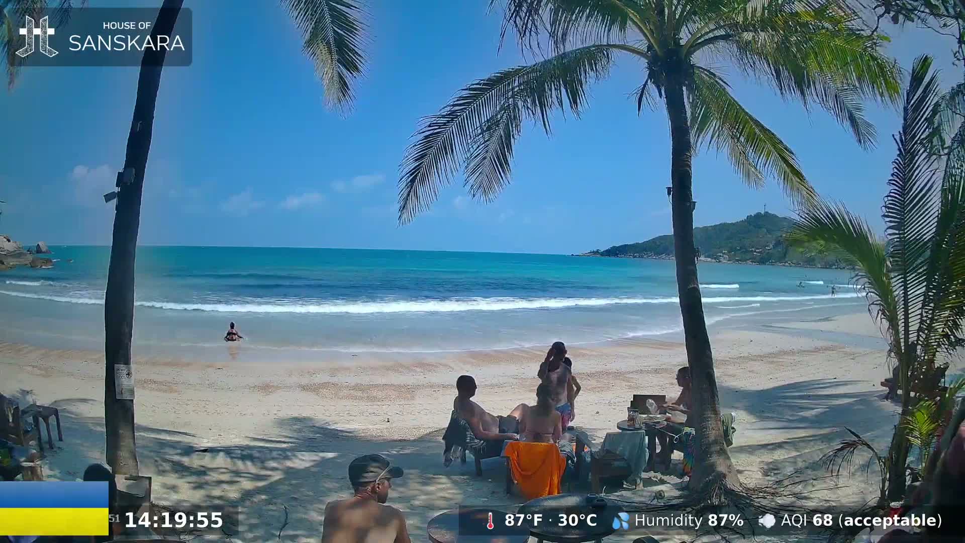 A sandy beach with turquoise water under a sunny sky is populated by palm trees and a small group of people relaxing near a table.