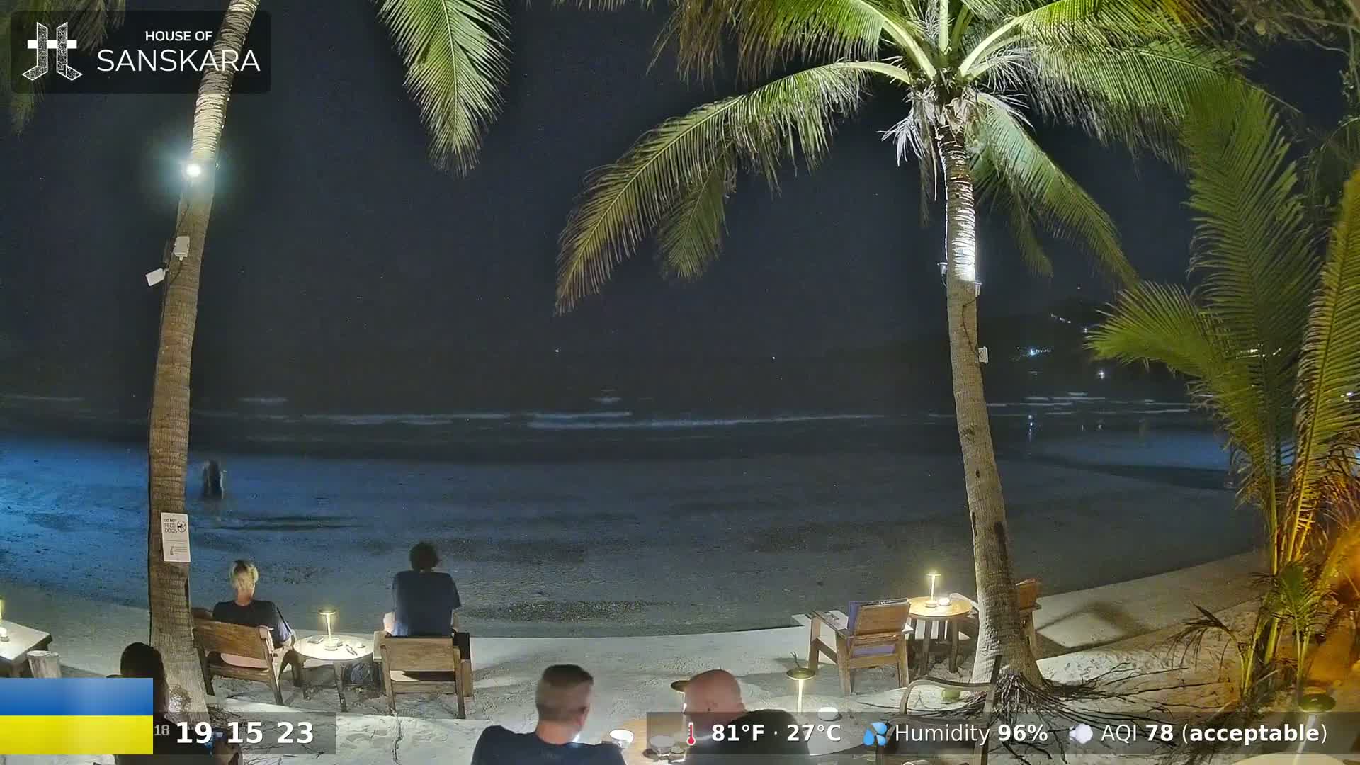 A nighttime beach scene shows a calm ocean under a dark sky, with several people sitting at tables under palm trees on the sand.