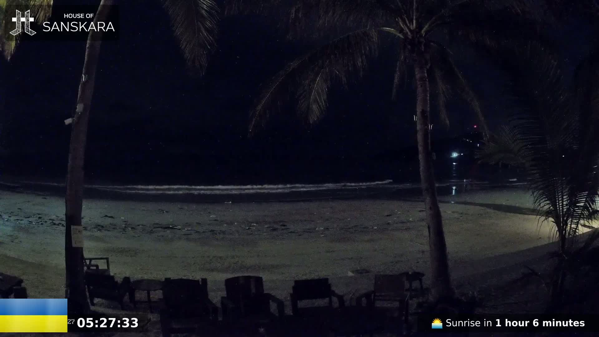 A dark, nighttime beach scene shows gently lapping waves under a starlit sky, palm trees in the foreground, and several empty chairs on the sand.