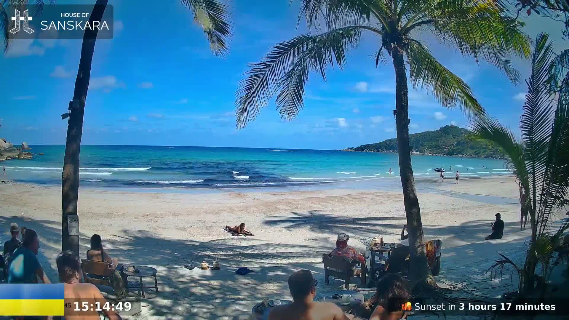 A sandy beach with turquoise water and gentle waves is partially shaded by palm trees, with several people relaxing on the sand and in beach chairs under a sunny sky.
