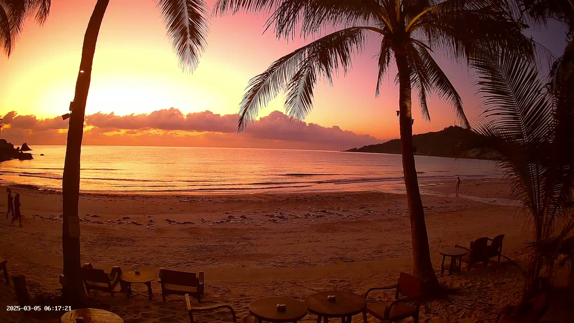 A sandy beach with palm trees and outdoor seating overlooks a calm ocean during a vibrant sunset.