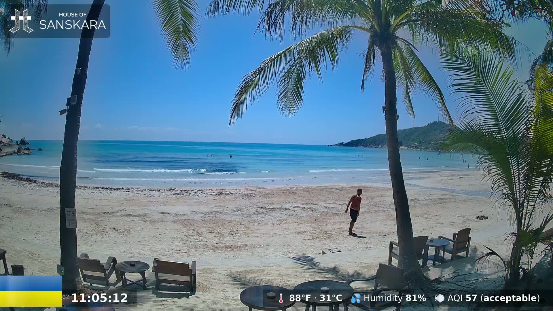 A sandy beach with clear turquoise water under a bright sunny sky, palm trees, and a person walking on the sand.