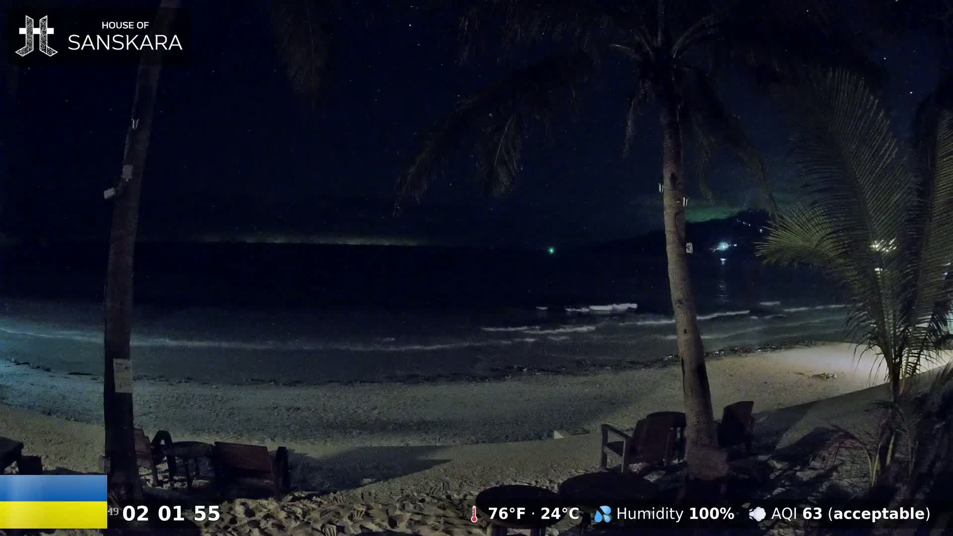 A nighttime beach scene shows gently lapping waves under a starry sky, palm trees silhouetted against the dark water, and empty outdoor seating near the shore.