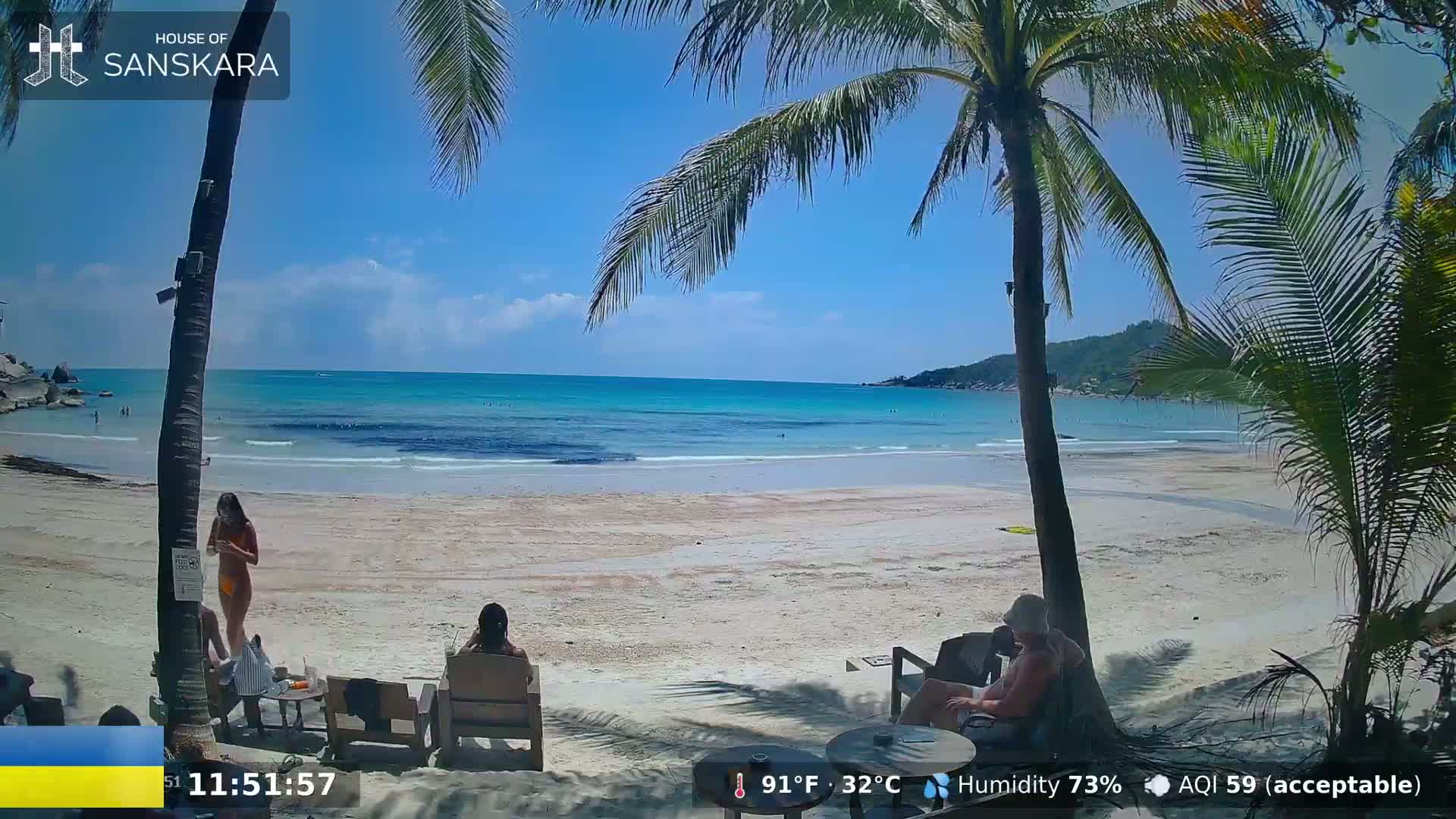 A sandy beach with calm turquoise water under a sunny sky is seen from under palm trees, with a few people relaxing on beach chairs.