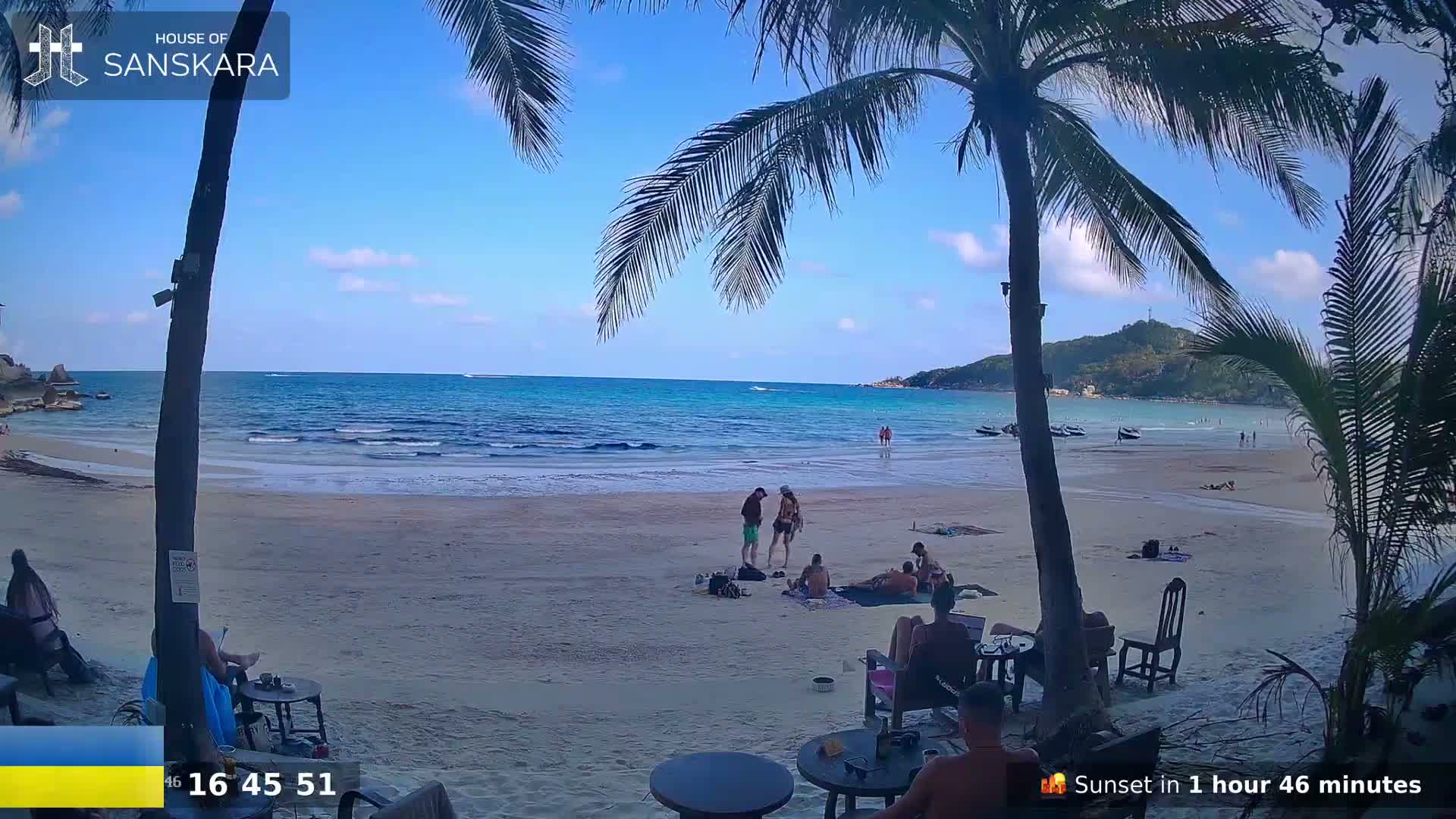A sandy beach with calm, blue ocean under a clear blue sky is viewed from behind palm trees, showing several people relaxing on the beach and at outdoor tables.
