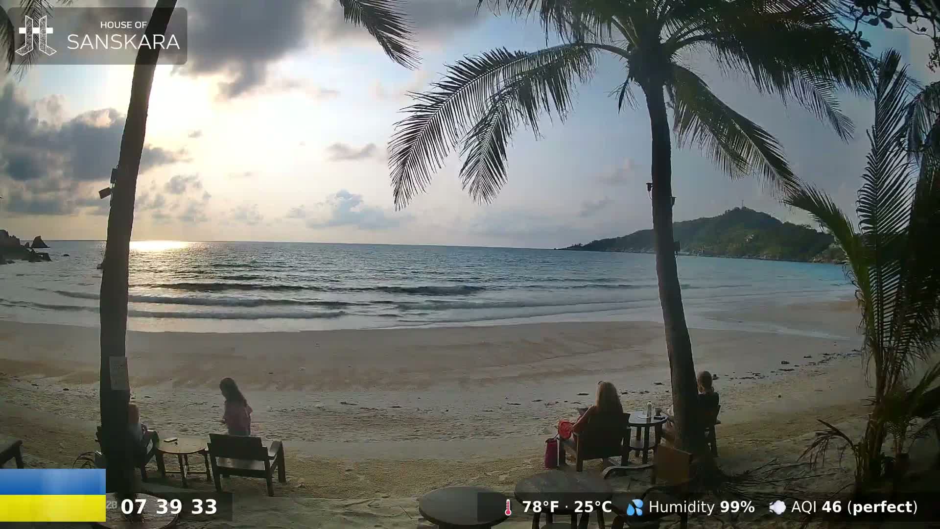A sandy beach with calm ocean waves and palm trees under a partly cloudy sky at sunset, with several people sitting at outdoor tables.