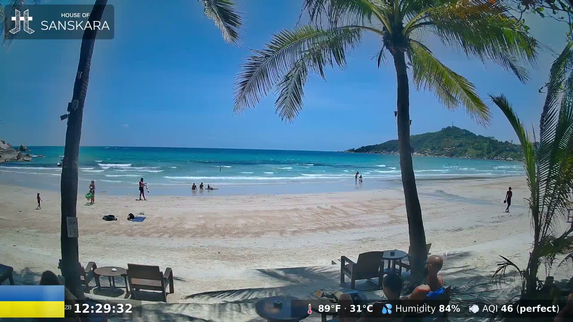 A sunny beach scene shows light-colored sand, calm turquoise water, palm trees, and several people relaxing or swimming.