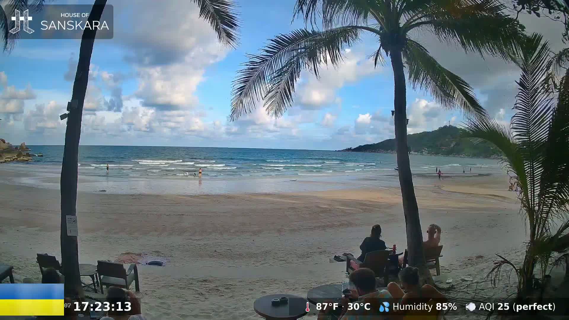 A sandy beach with calm ocean waves, partly cloudy skies, and people relaxing under palm trees.