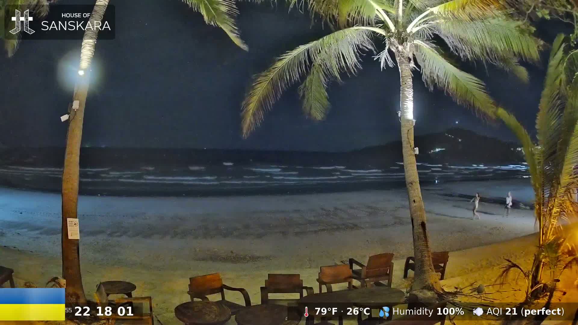 A nighttime beach scene shows a calm ocean with gentle waves under a clear, dark sky, two palm trees with lights illuminating the area, and several empty wooden chairs and tables on a sandy beach.