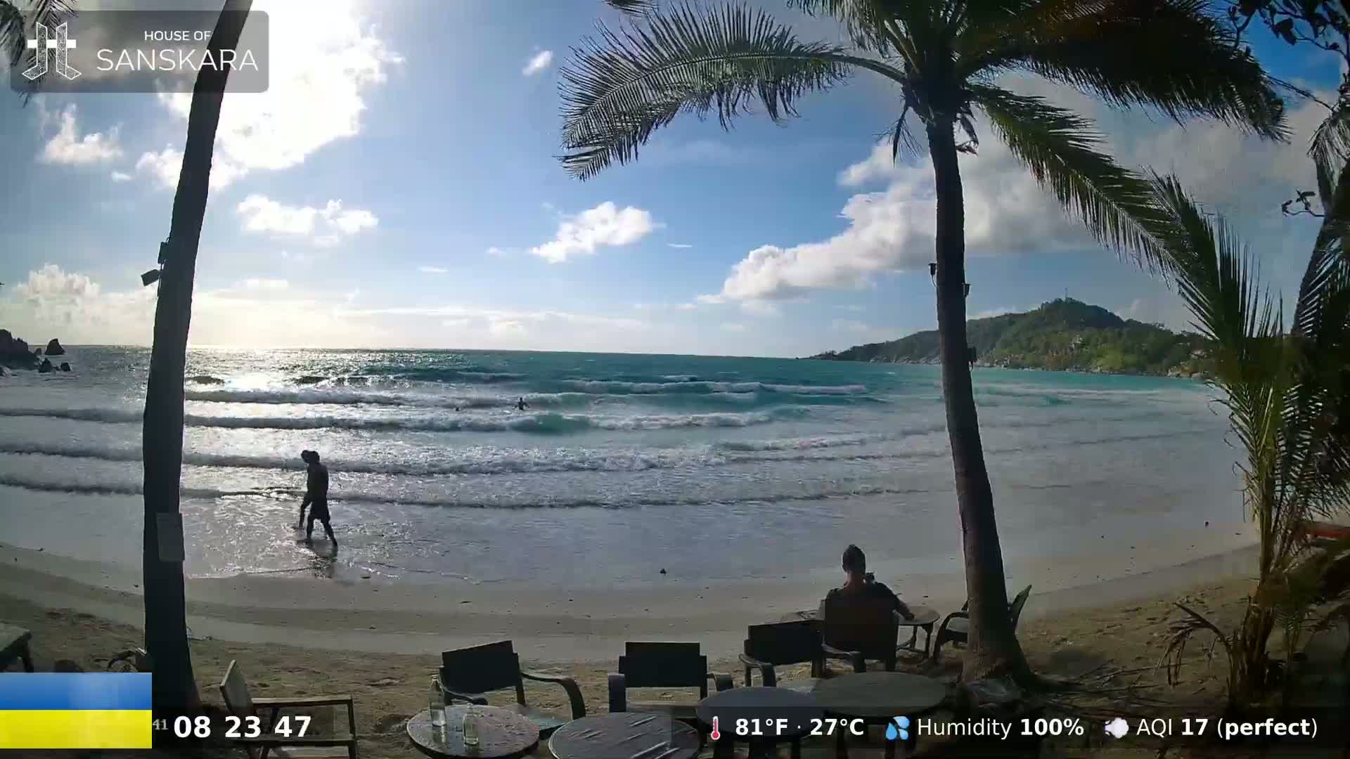 A sunny beach scene shows a person wading in the ocean's gentle waves, another person sitting at an outdoor table, and palm trees framing a view of the water and a distant hill under a partly cloudy sky.
