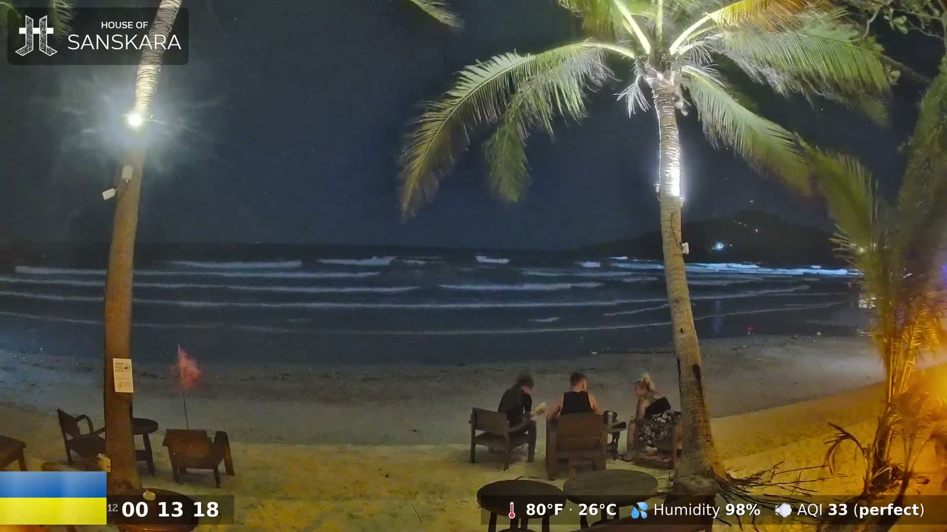 At night, under a clear, starry sky, several people sit at tables on a sandy beach beside gently lapping waves, illuminated by outdoor lighting near palm trees.