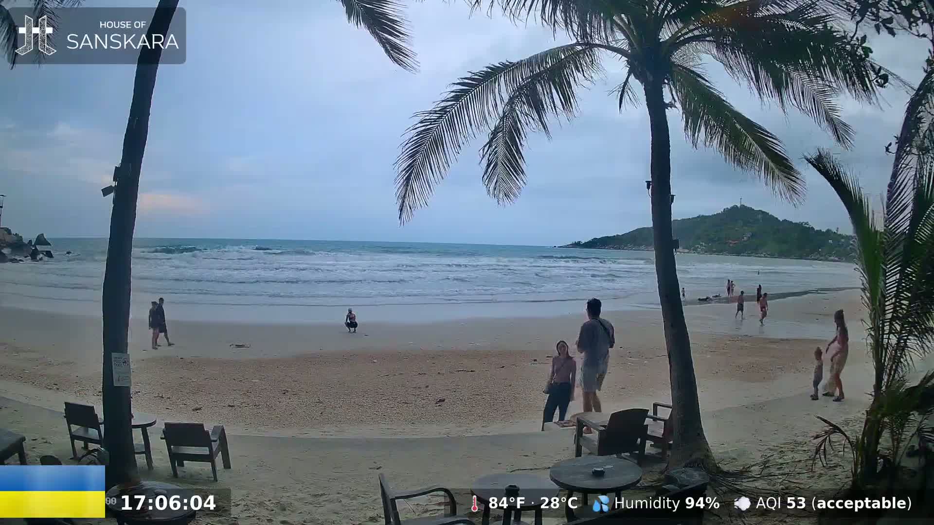 A sandy beach with a few people walking and playing in the shallow water under an overcast sky.