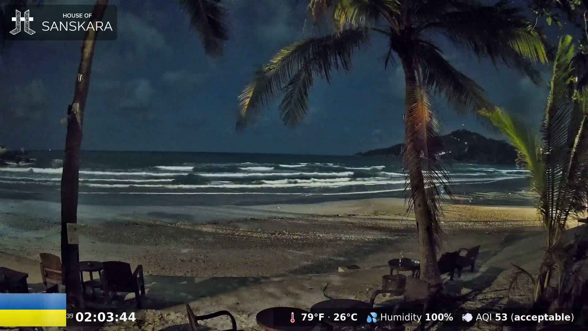 A nighttime beach scene shows gentle waves lapping a dark sandy shore under a partly cloudy sky, with palm trees in the foreground and a hill in the distance.