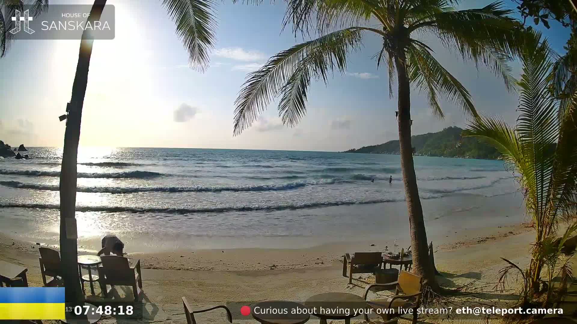 A sandy beach with calm waves, palm trees, and outdoor furniture is shown under a partly sunny sky.