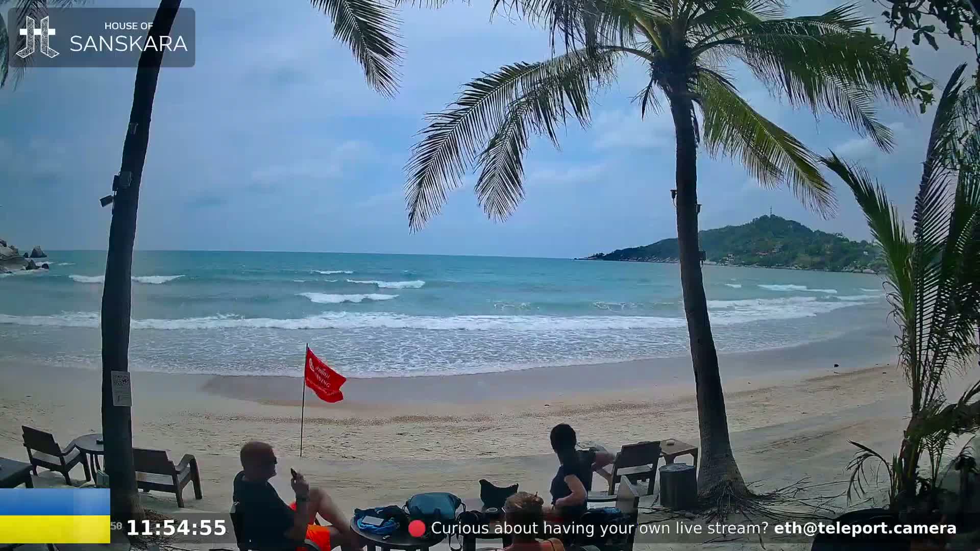 A sandy beach with gentle waves, palm trees, and several people sitting under the shade, under partly cloudy skies.