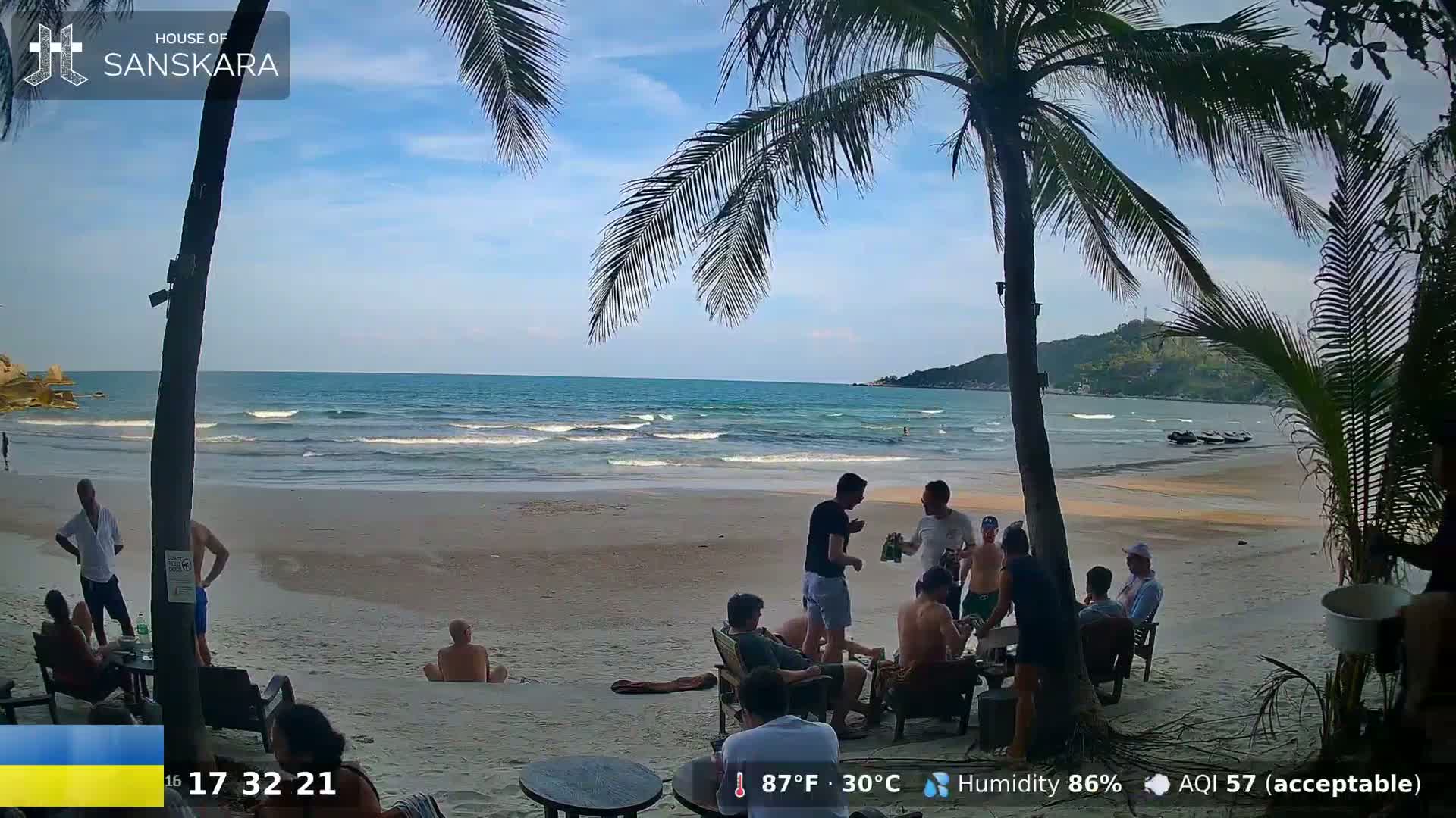 A sandy beach with palm trees and a group of people sitting and standing on the shore, under a partly sunny sky, and a calm ocean in the background.