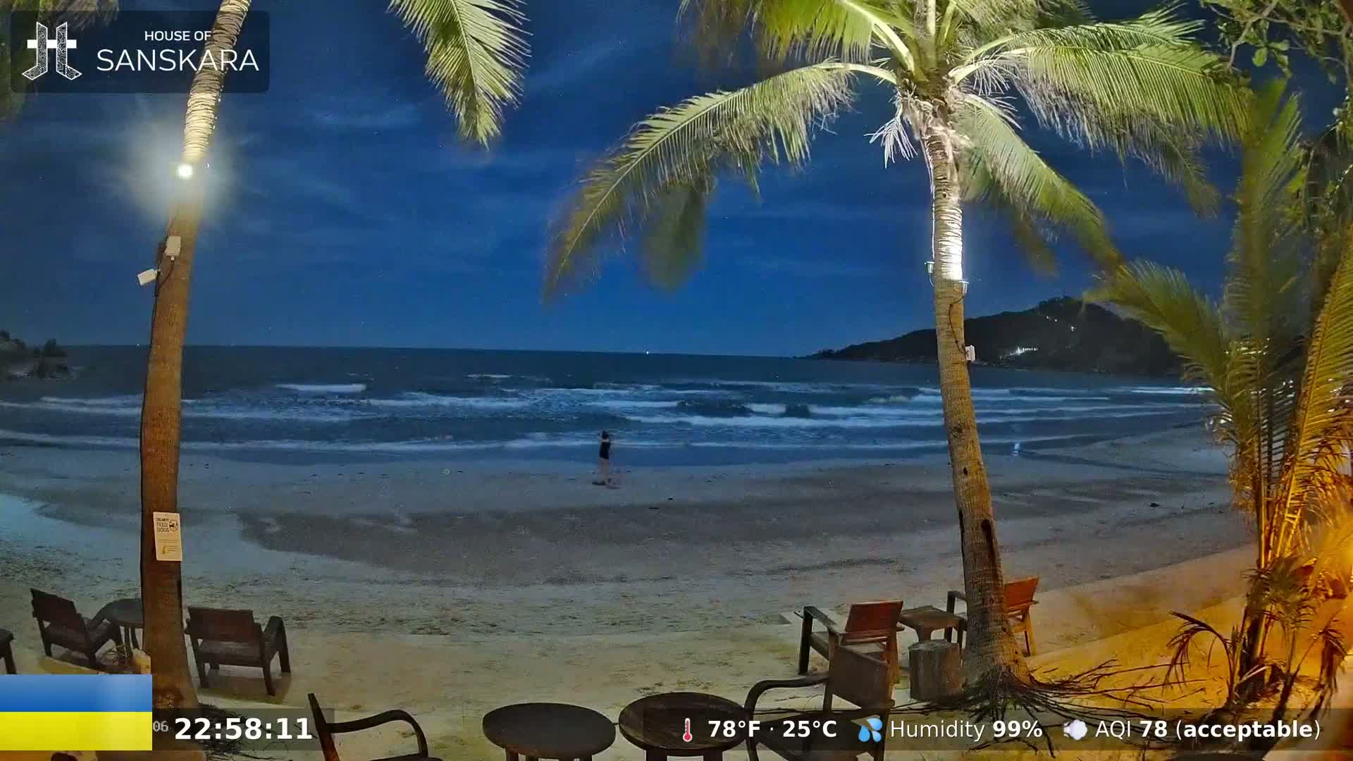 A nighttime beach scene shows a person walking along the shore under a mostly clear, dark blue sky, with palm trees, and outdoor furniture nearby.