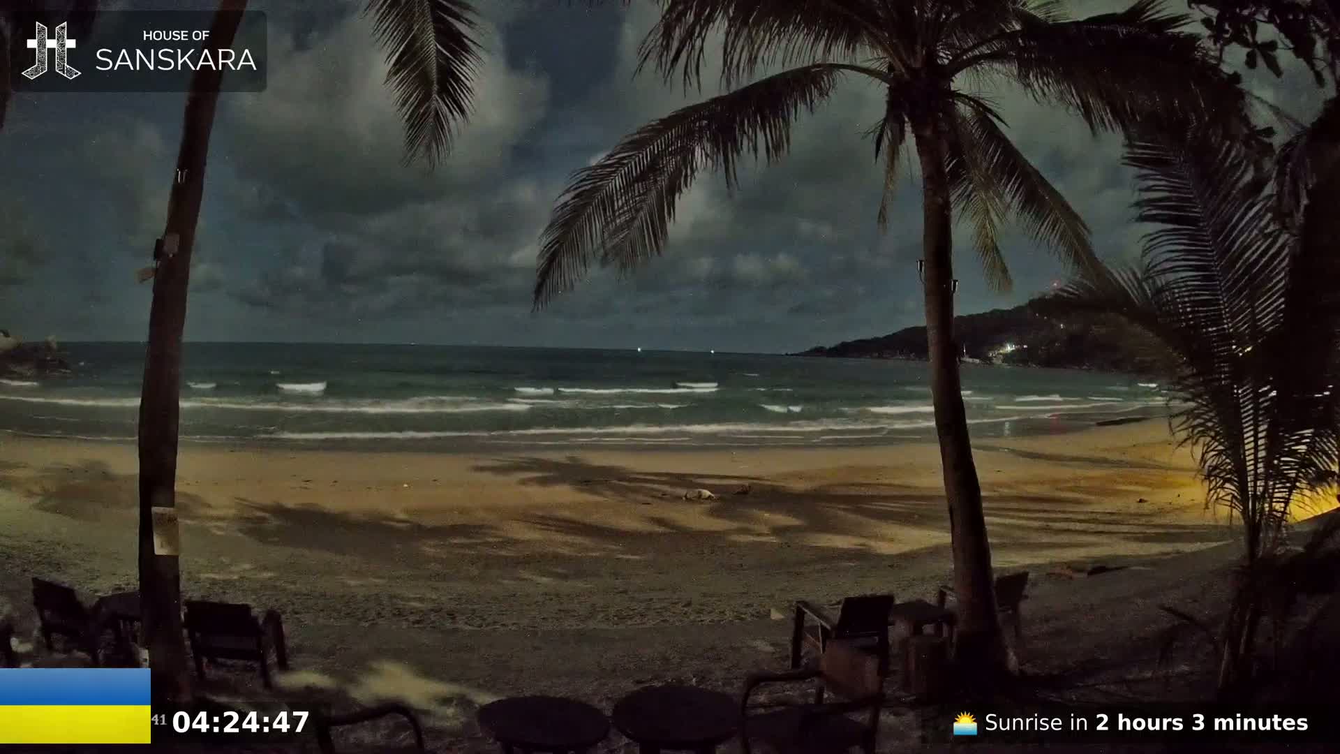 Under a partly cloudy night sky, a dark sandy beach is gently lapped by waves, with palm trees in the foreground and a few dark chairs and tables.