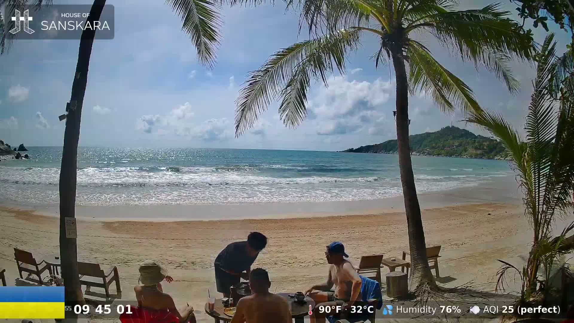 On a sunny day with a slight breeze, several people sit at an outdoor table on a sandy beach next to a calm ocean, partially shaded by palm trees.