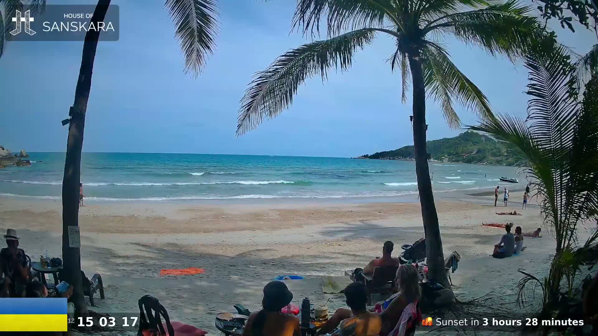 A sandy beach with calm waves, some people relaxing, and palm trees framing a view of the ocean under a clear blue sky.