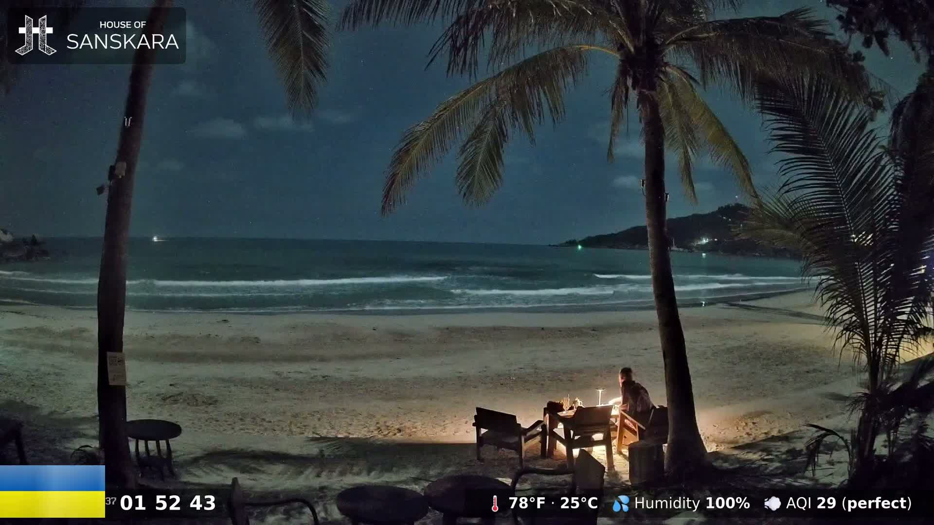 A person sits at an outdoor table on a sandy beach at night, illuminated by a small fire, under palm trees, with calm waves lapping the shore under a dark sky.