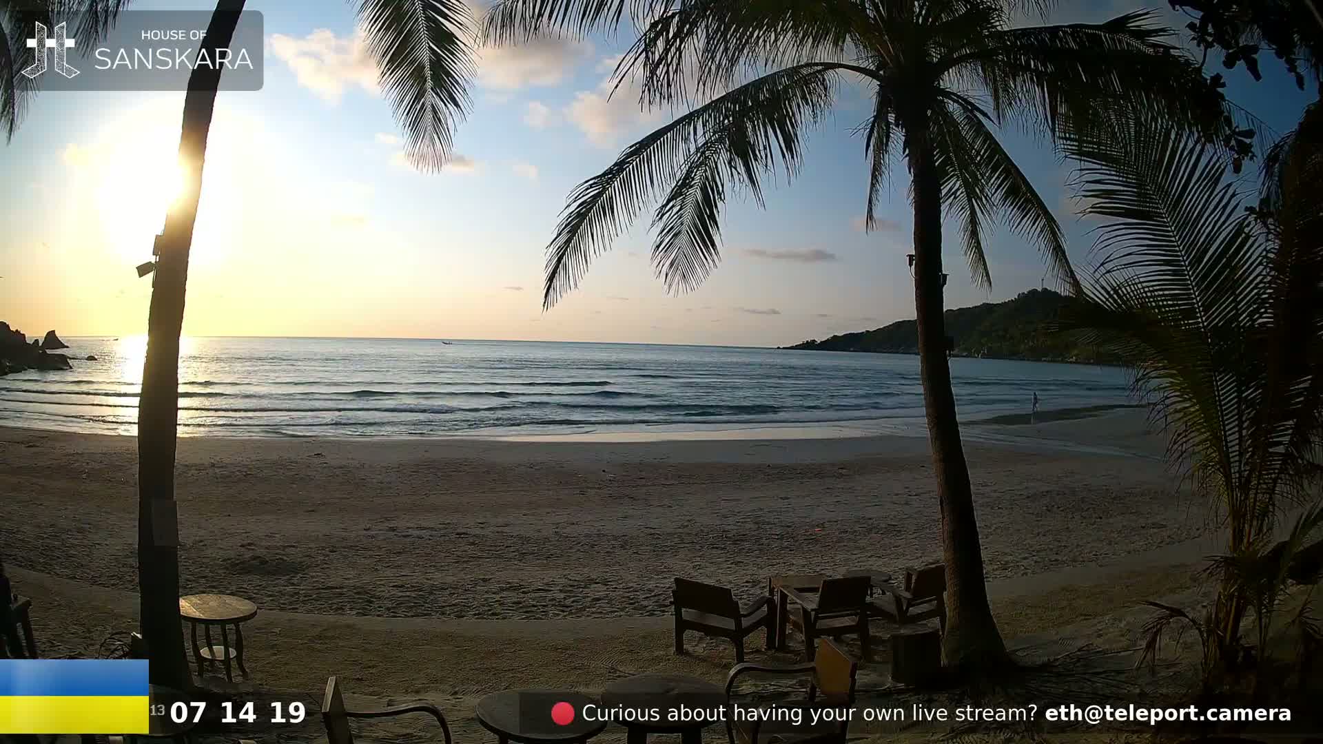 A beach scene at sunset shows calm ocean waves lapping a sandy shore, palm trees in the foreground, and outdoor furniture on the sand.