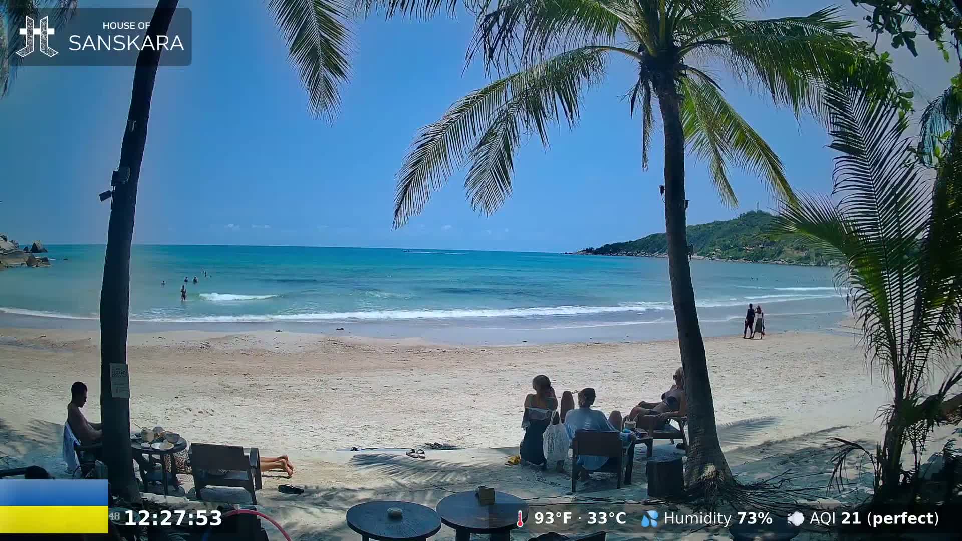 A sandy beach with palm trees and a few people on it is shown, under a clear blue sky on a sunny day.