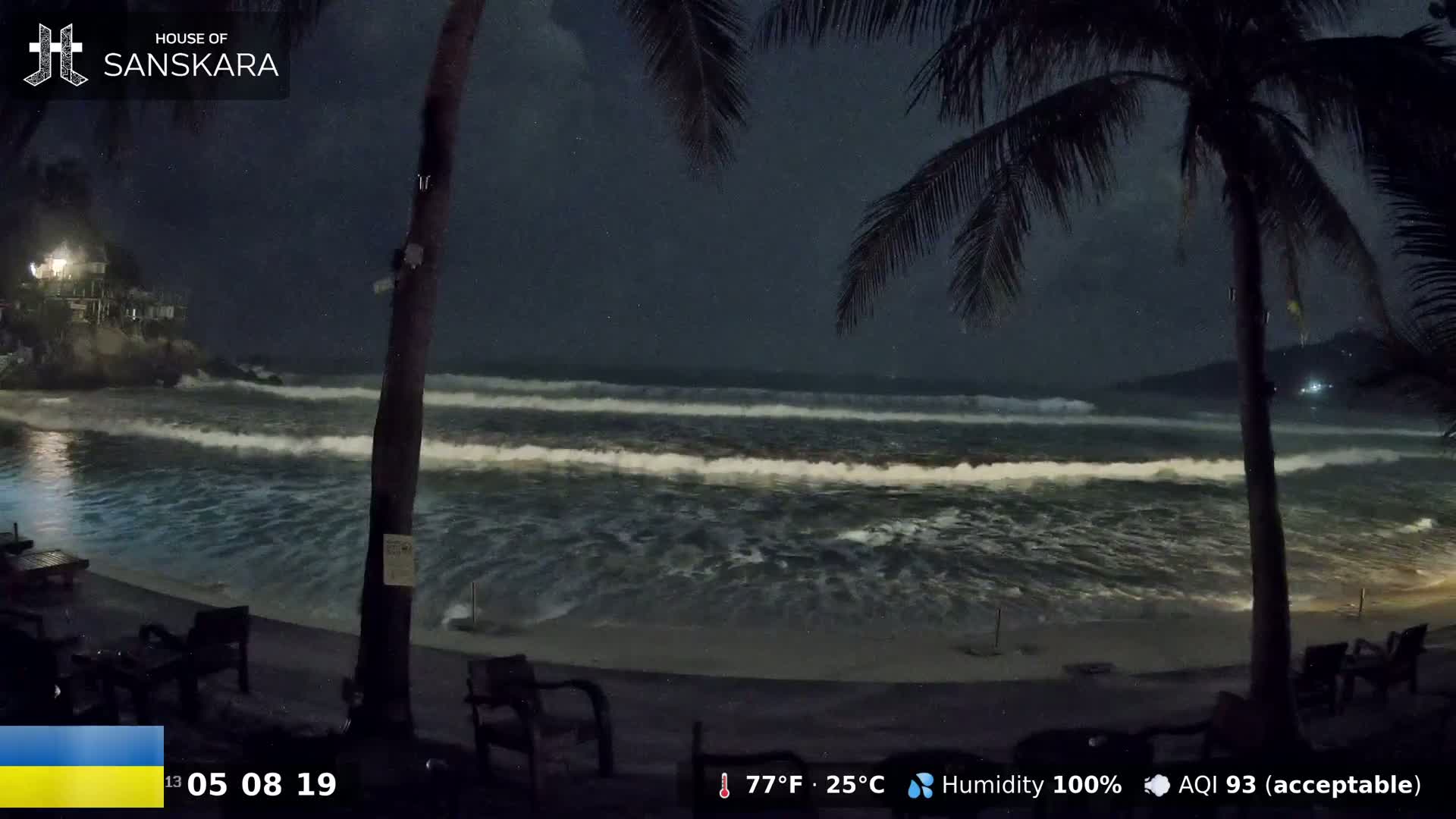 At night, under a dark sky with 100% humidity, moderate waves crash on a beach near palm trees and outdoor seating.