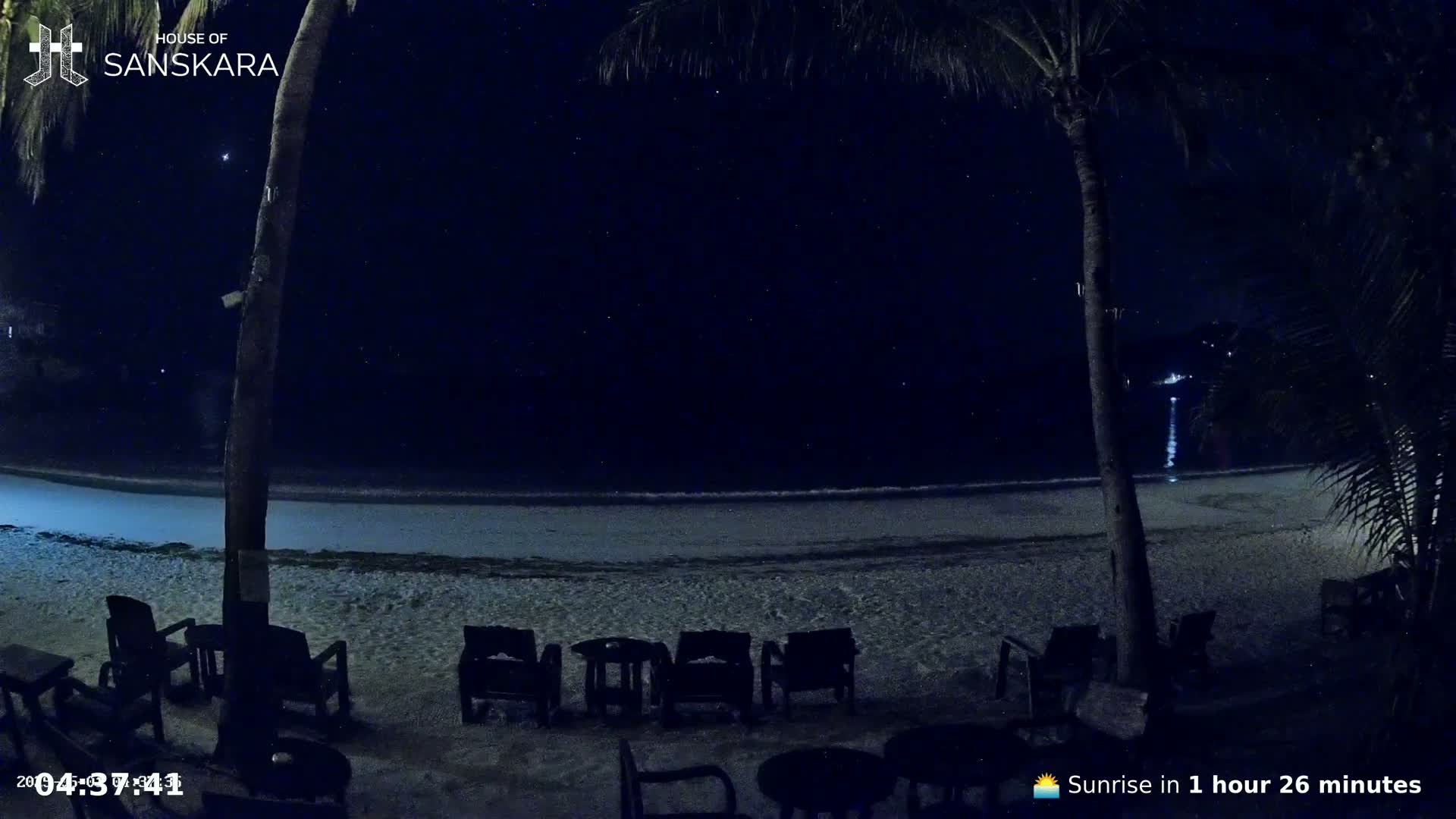 Empty chairs sit on a sandy beach at night under a starry sky, overlooking a calm ocean.