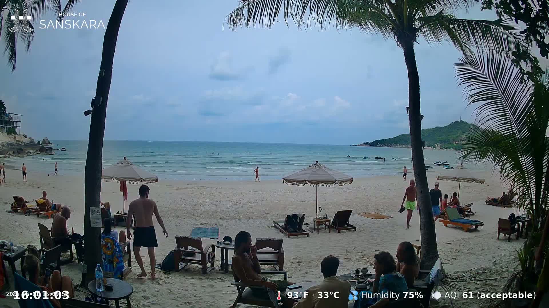 A sandy beach with calm ocean waves, palm trees, and beachgoers under a partly cloudy sky.