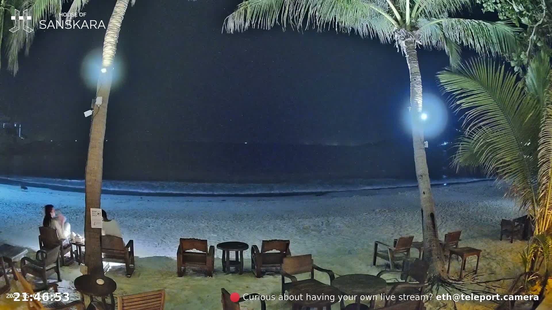 A nighttime beach scene shows a sandy shore with gentle waves lapping at the edge, palm trees, and outdoor seating at a beachfront establishment.