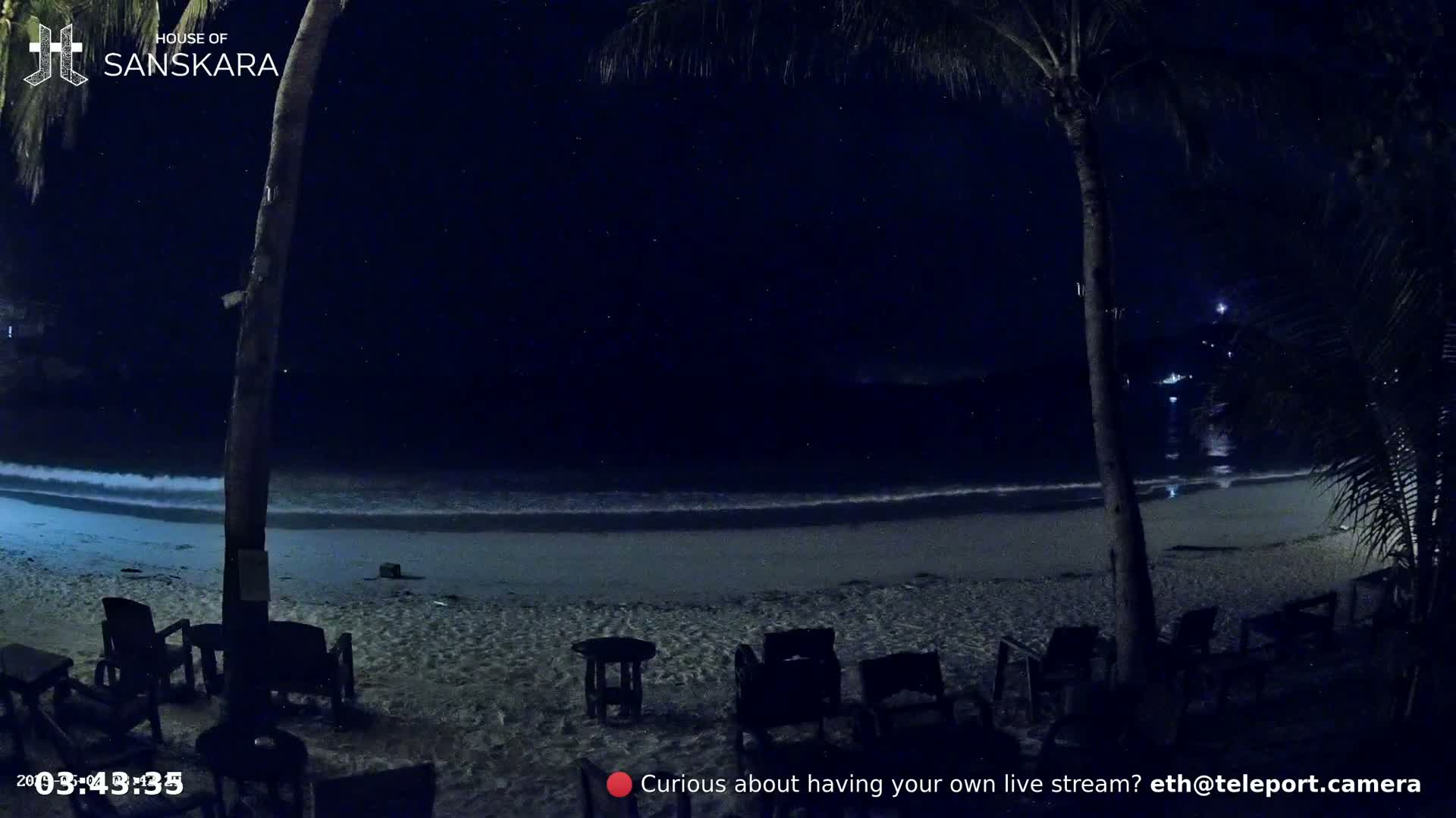 A nighttime beach scene shows a sandy shore with gentle waves lapping under a clear, starlit sky, palm trees and several empty chairs are visible.