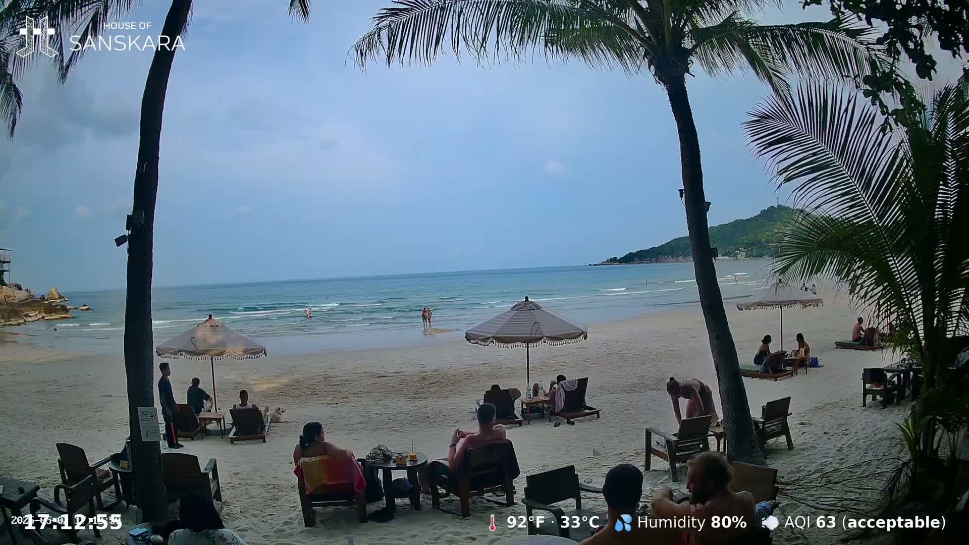 A sandy beach with calm ocean waves, palm trees, and people relaxing under umbrellas on a partly cloudy day.