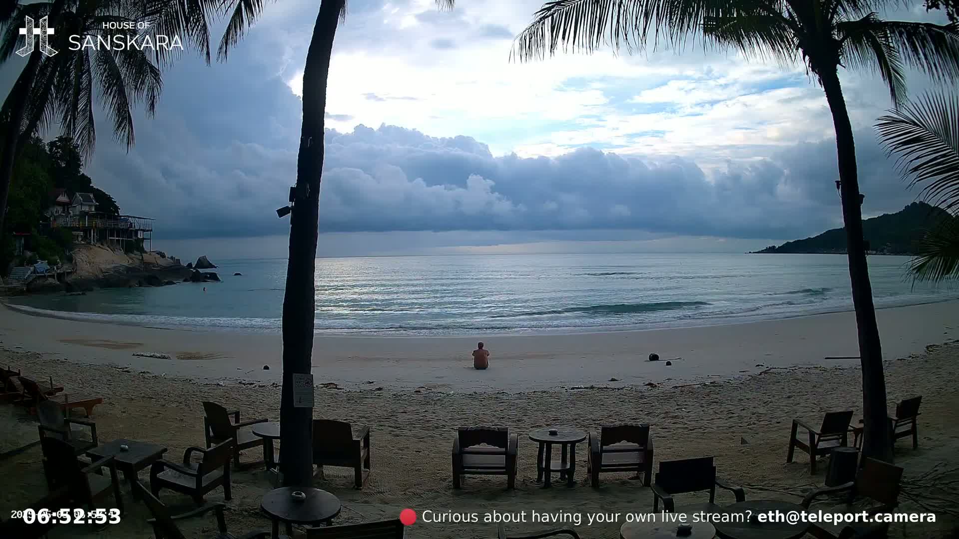 A person sits on a sandy beach, facing a calm ocean under a cloudy sky, with palm trees and outdoor seating visible in the foreground.