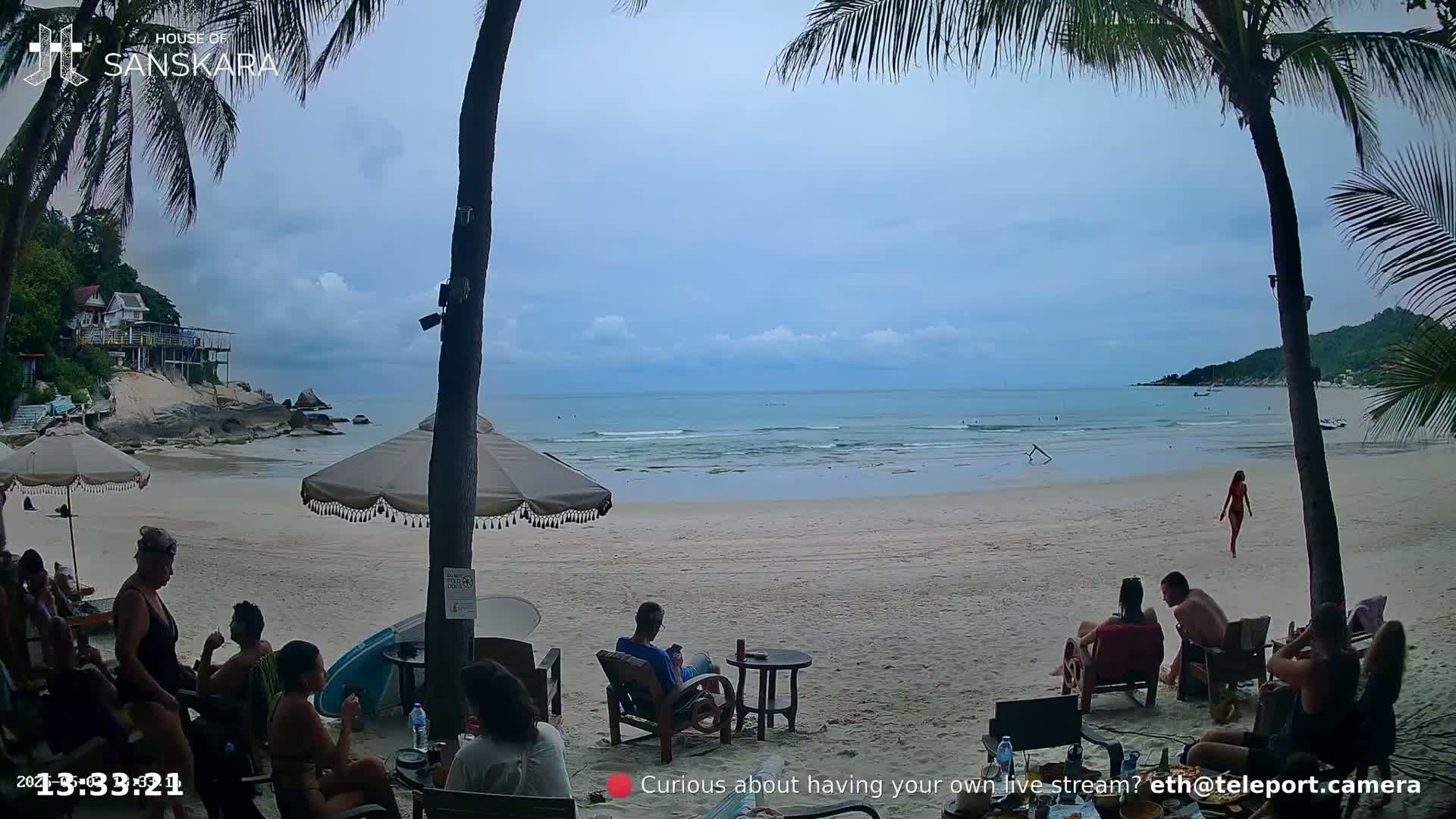 A sandy beach with calm ocean waves, palm trees, and several people relaxing under umbrellas on a partly cloudy day.