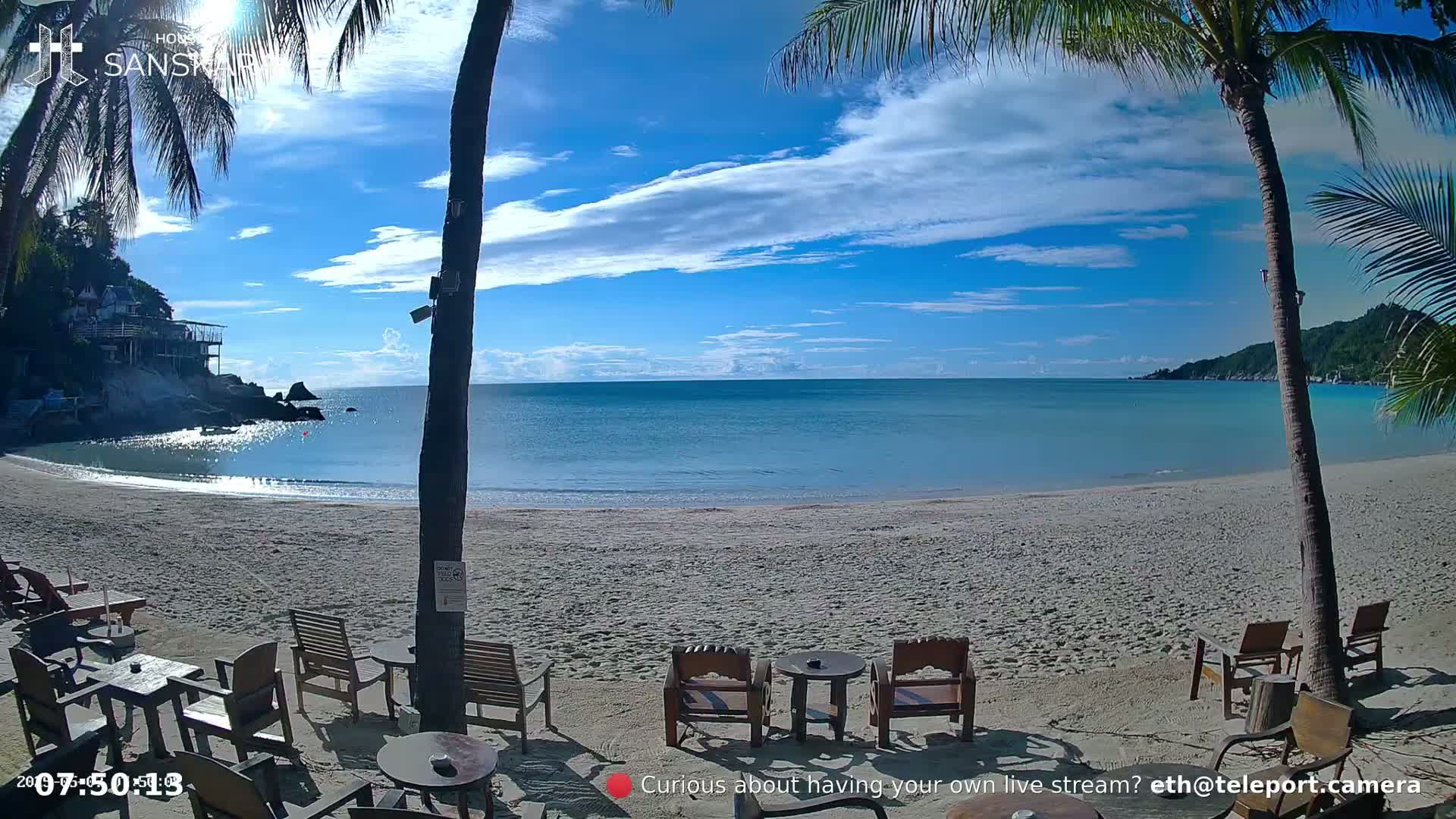A sandy beach with calm, turquoise water and a few empty wooden chairs and tables under palm trees on a partly cloudy day.