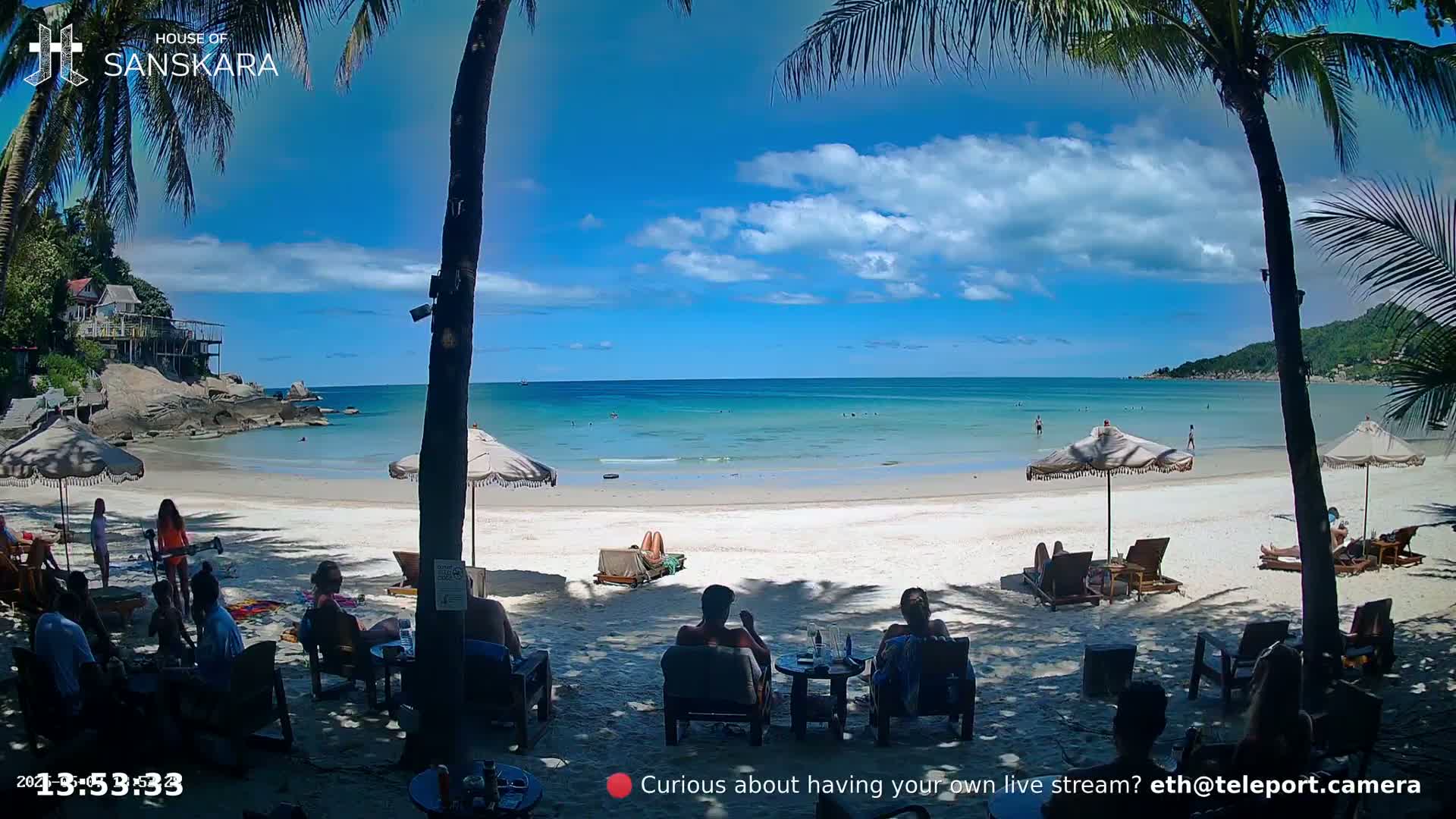 A sunny beach scene shows a calm turquoise ocean, white sand, palm trees, beach umbrellas, and people relaxing on lounge chairs.