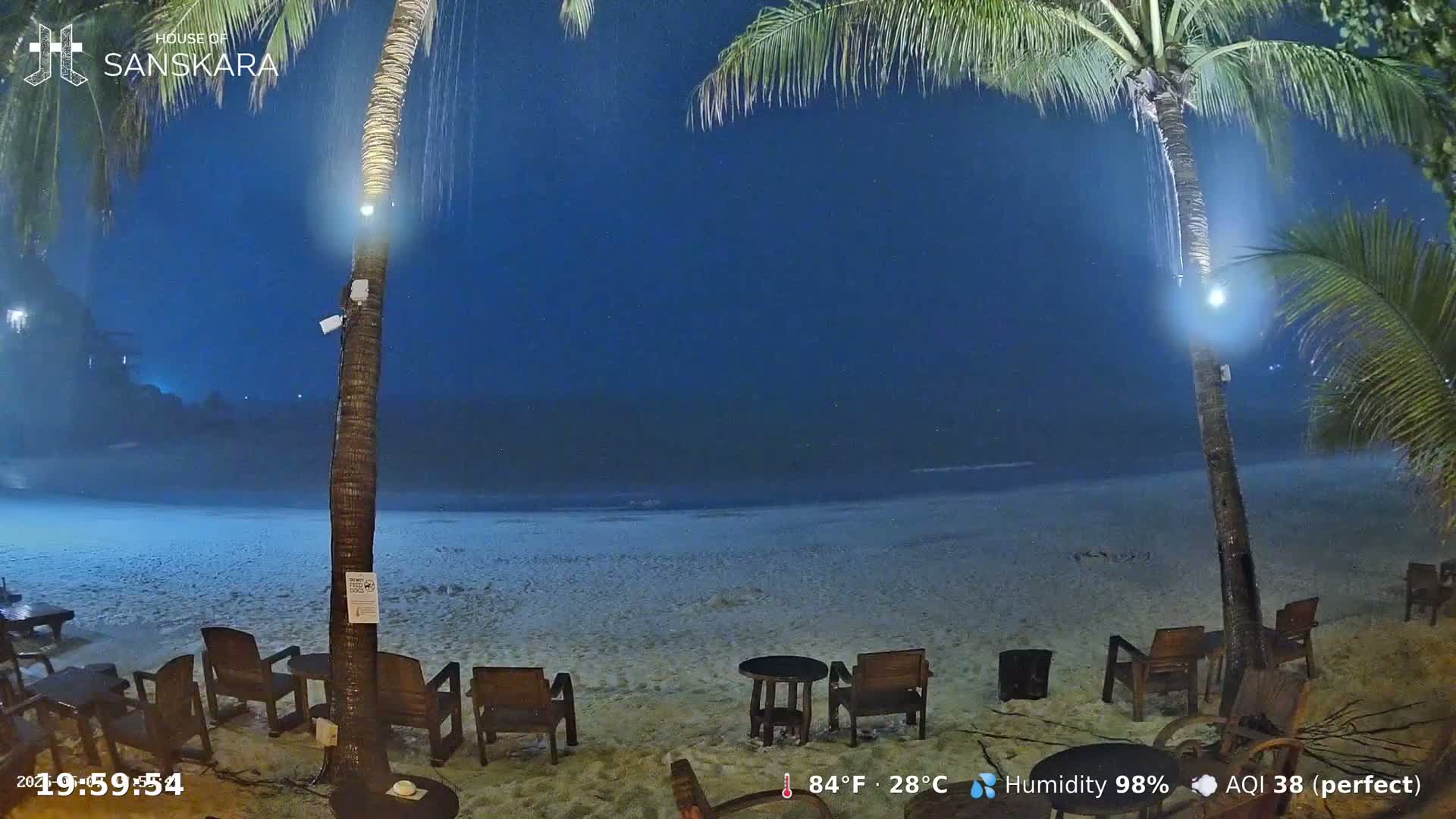 Empty wooden chairs and tables sit on a sandy beach at night during a light rain shower under a dark blue sky, illuminated by lights on palm trees.