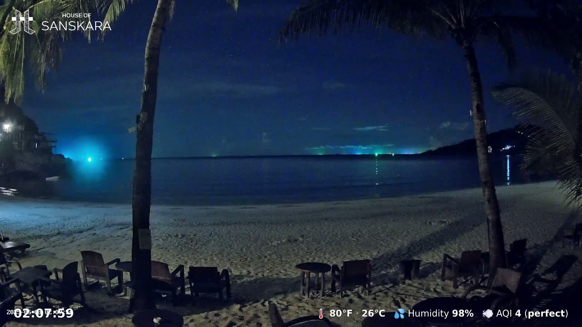 A dark, starry night at a beach with palm trees, empty outdoor seating, and a calm ocean.