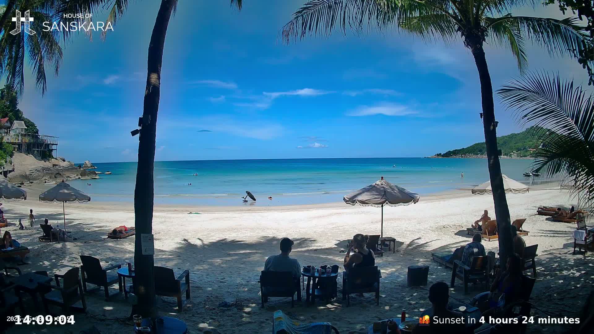 A sunny beach scene shows calm, teal water, white sand, palm trees, beach umbrellas, and people relaxing in chairs under the shade.