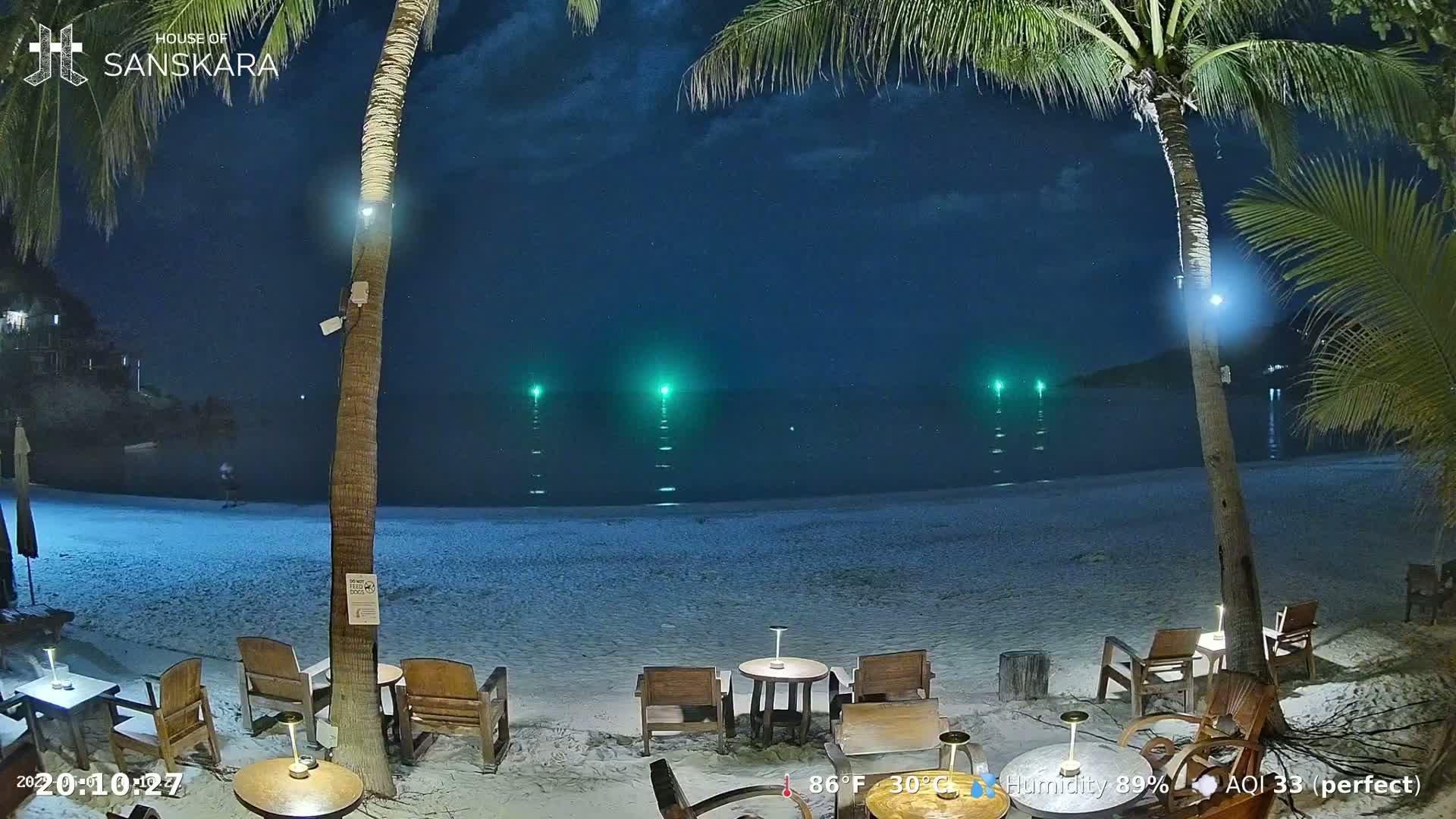 A nighttime beach scene with several empty tables and chairs under palm trees, a calm sea with several green lights in the distance, and a clear, starry sky.