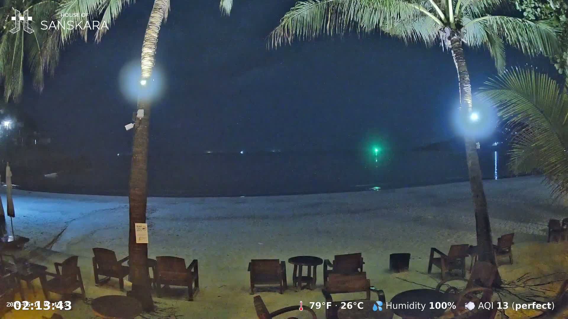 Empty chairs sit on a sandy beach at night under palm trees, with calm water visible in the background.