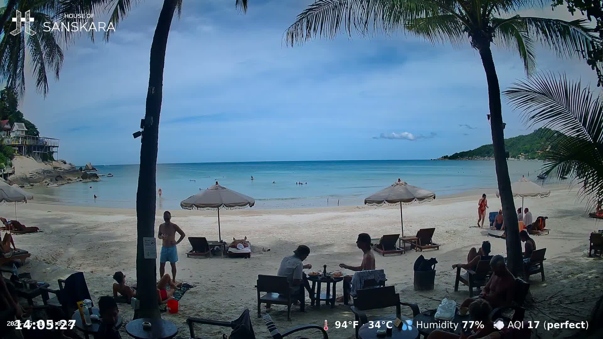 A beach scene shows people relaxing under umbrellas on a sandy beach with calm, clear turquoise water, palm trees, and a partly sunny sky.