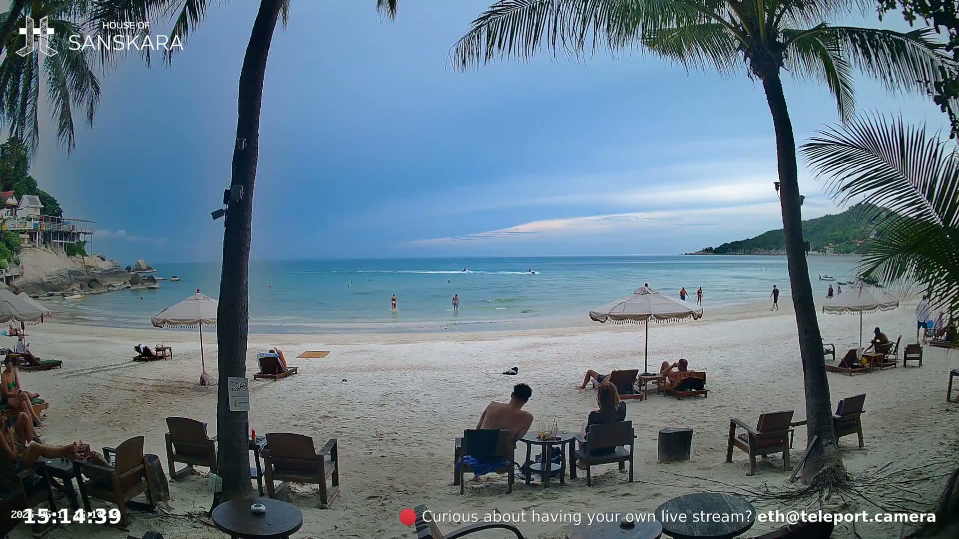 A sandy beach with palm trees, people relaxing on chairs and in the water, and calm, light-blue ocean under a partly cloudy sky.