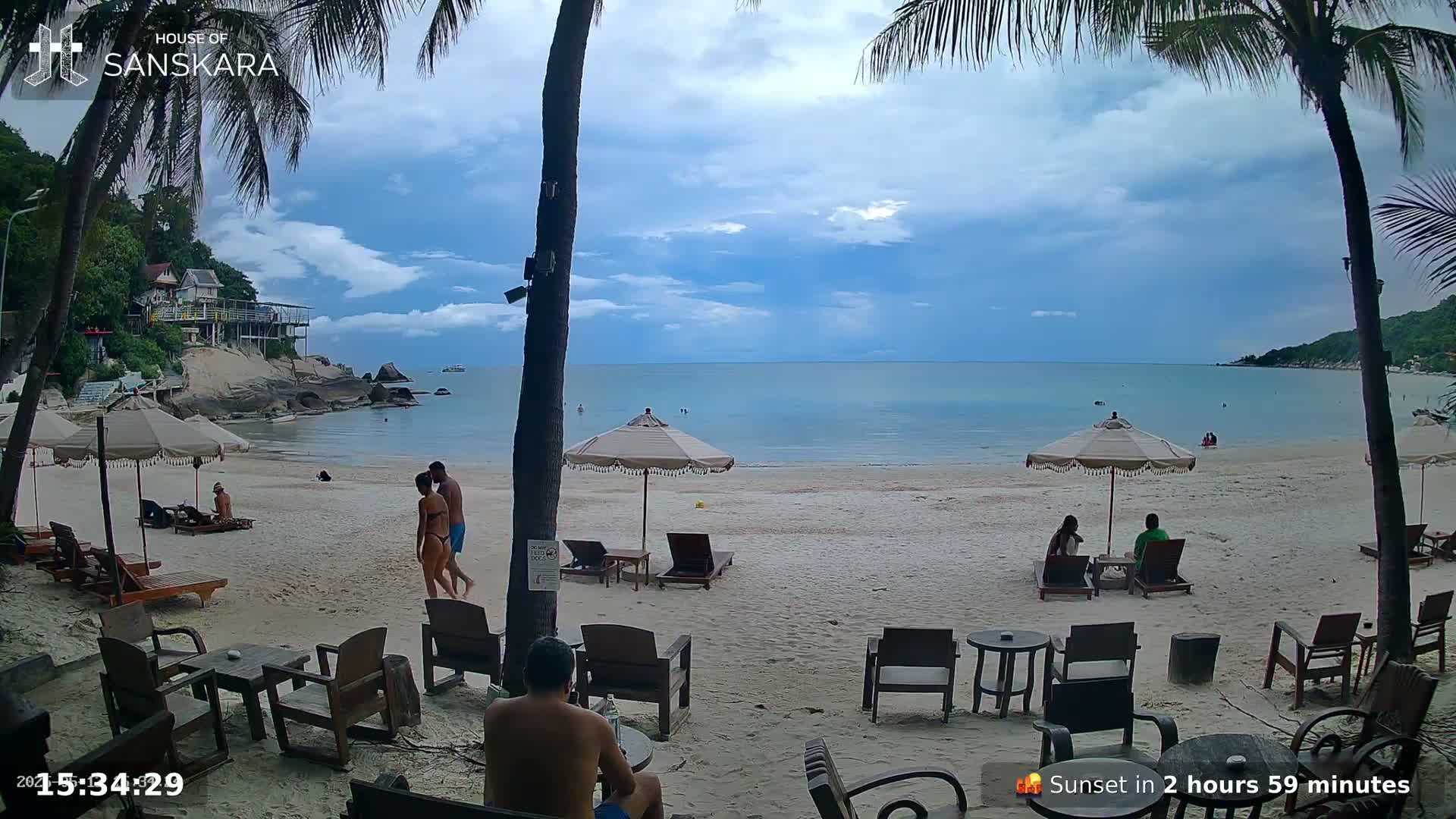 A partly cloudy day at a beach with palm trees, beach chairs and umbrellas, and several people relaxing.