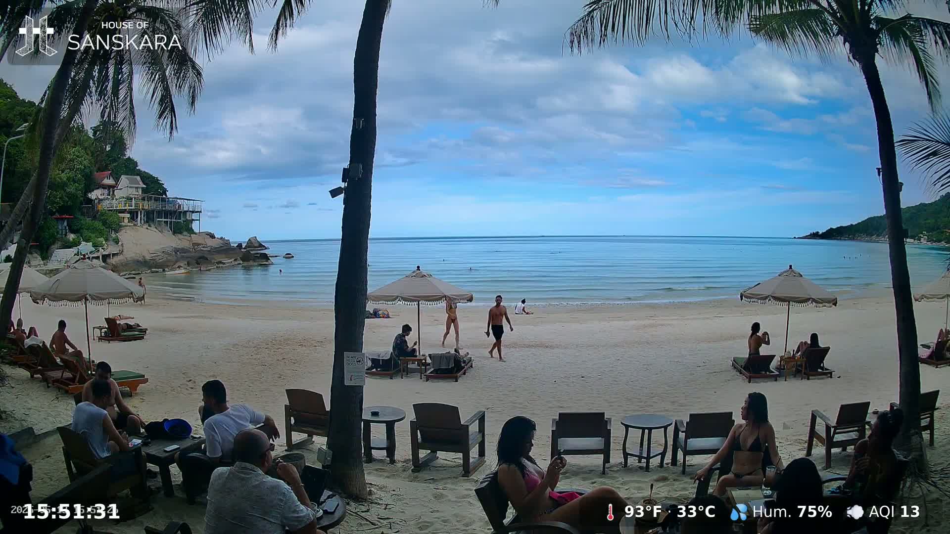 A partly cloudy day at a beachside café, where people relax under umbrellas on a sandy beach with calm, light-blue water.