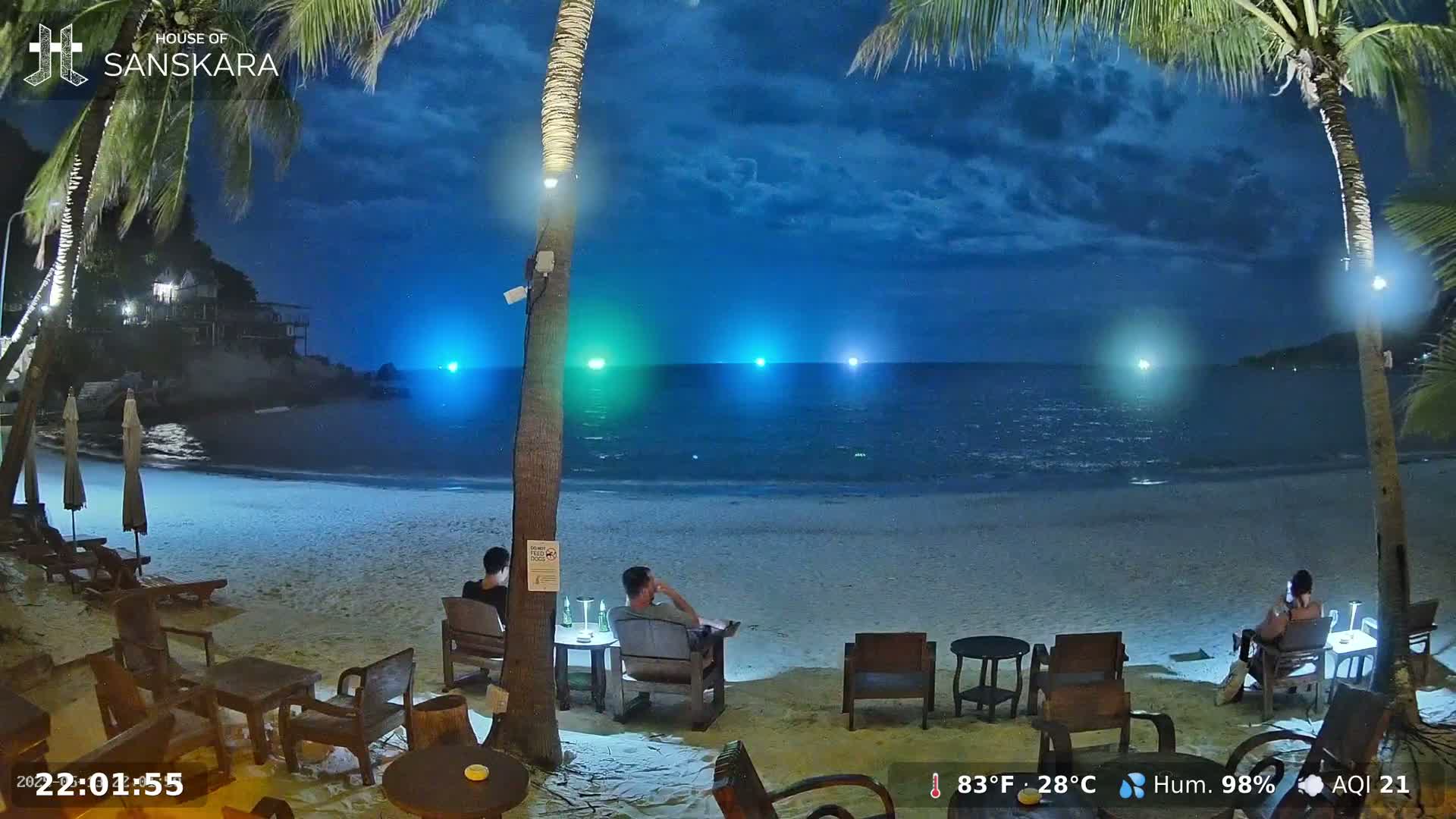 A nighttime beach scene shows several people seated at outdoor tables under palm trees, with a calm sea and a partly cloudy sky.