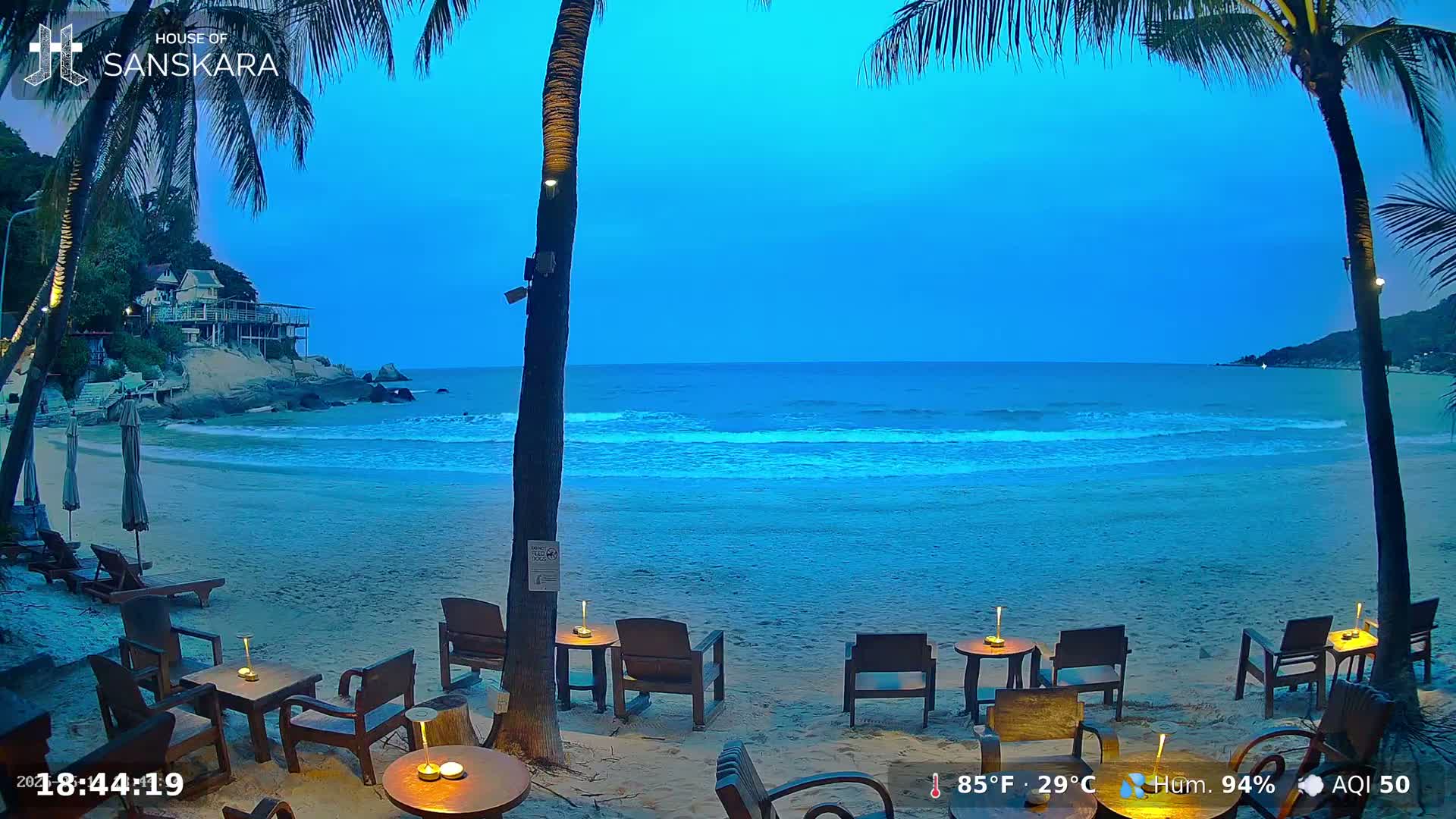 A sandy beach at twilight with calm ocean waves, palm trees, and outdoor seating areas with small tables and chairs.