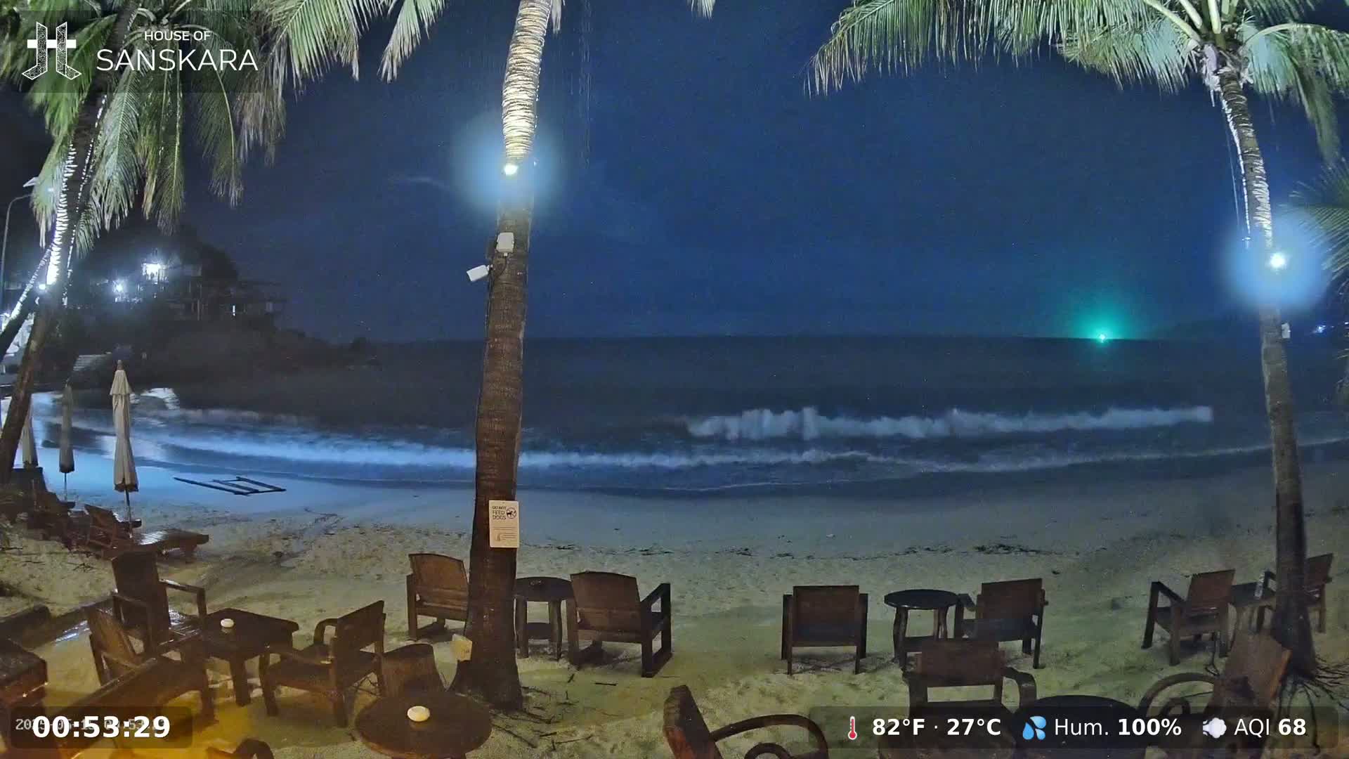 A nighttime beach scene shows gently lapping waves under a dark blue, clear sky, with palm trees and outdoor furniture illuminated near the shore.